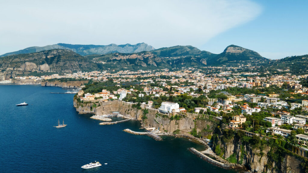 Aerial drone view of the Tyrrhenian sea coast in Sorrento, Italy. Multiple classic buildings on rocky cliffs, a lot of greenery and hills in the distance